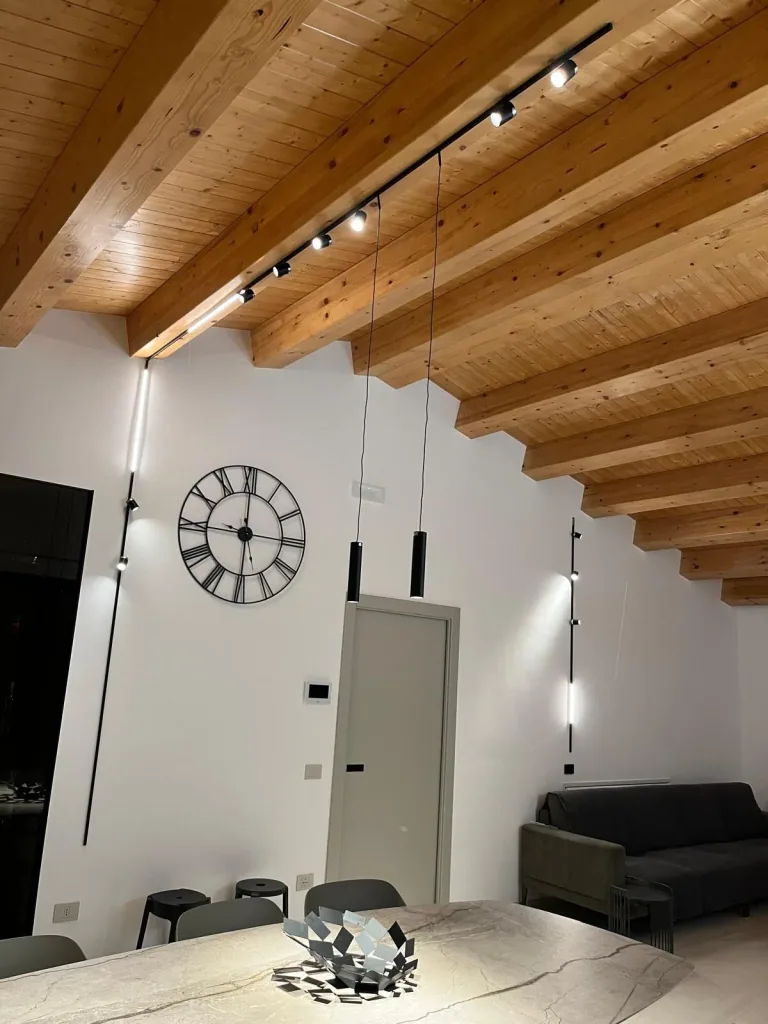 Soggiorno con soffitto in legno a vista, binario sottile e sospensioni lineari accese